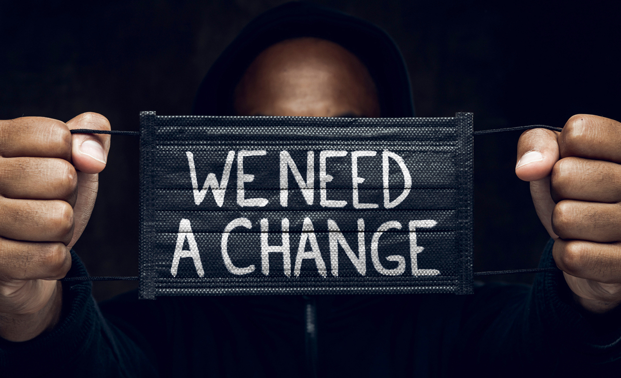 Black man holding a medical facemask with "We Need A Change" written on it.