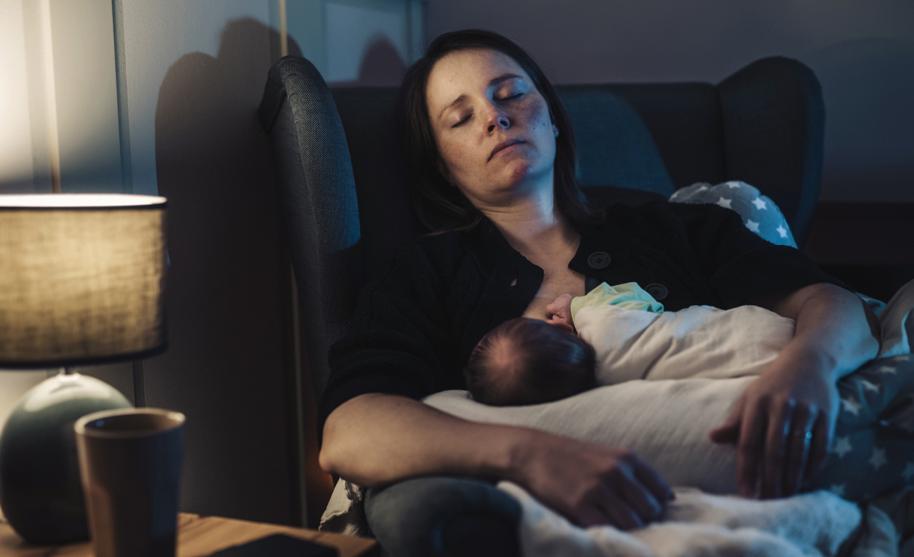 An exhausted mother breast feeding her baby in somber lighting.