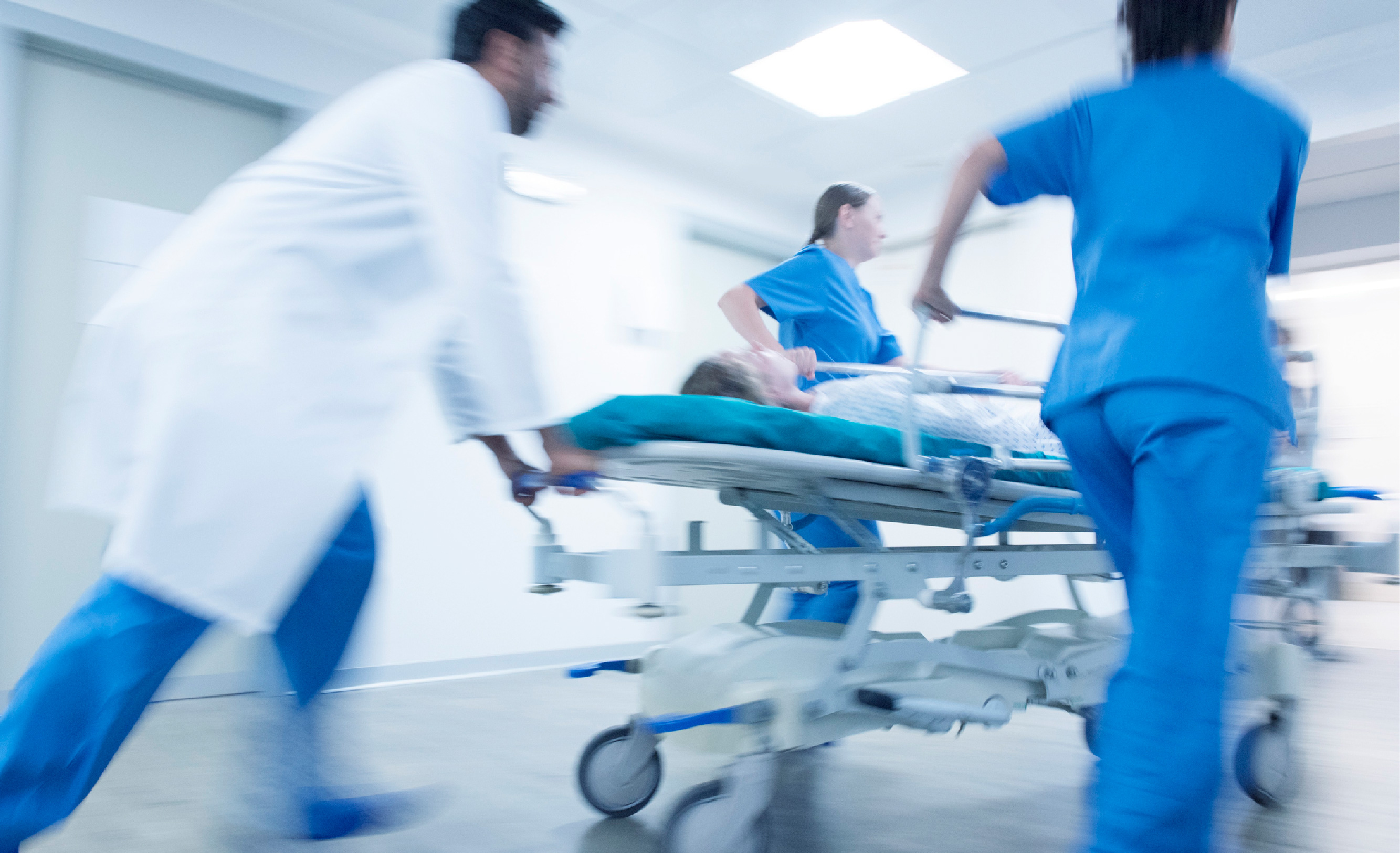 Healthcare workers in scrubs and one in a white coat rush an individual in a hospital gown through a hospital hallway