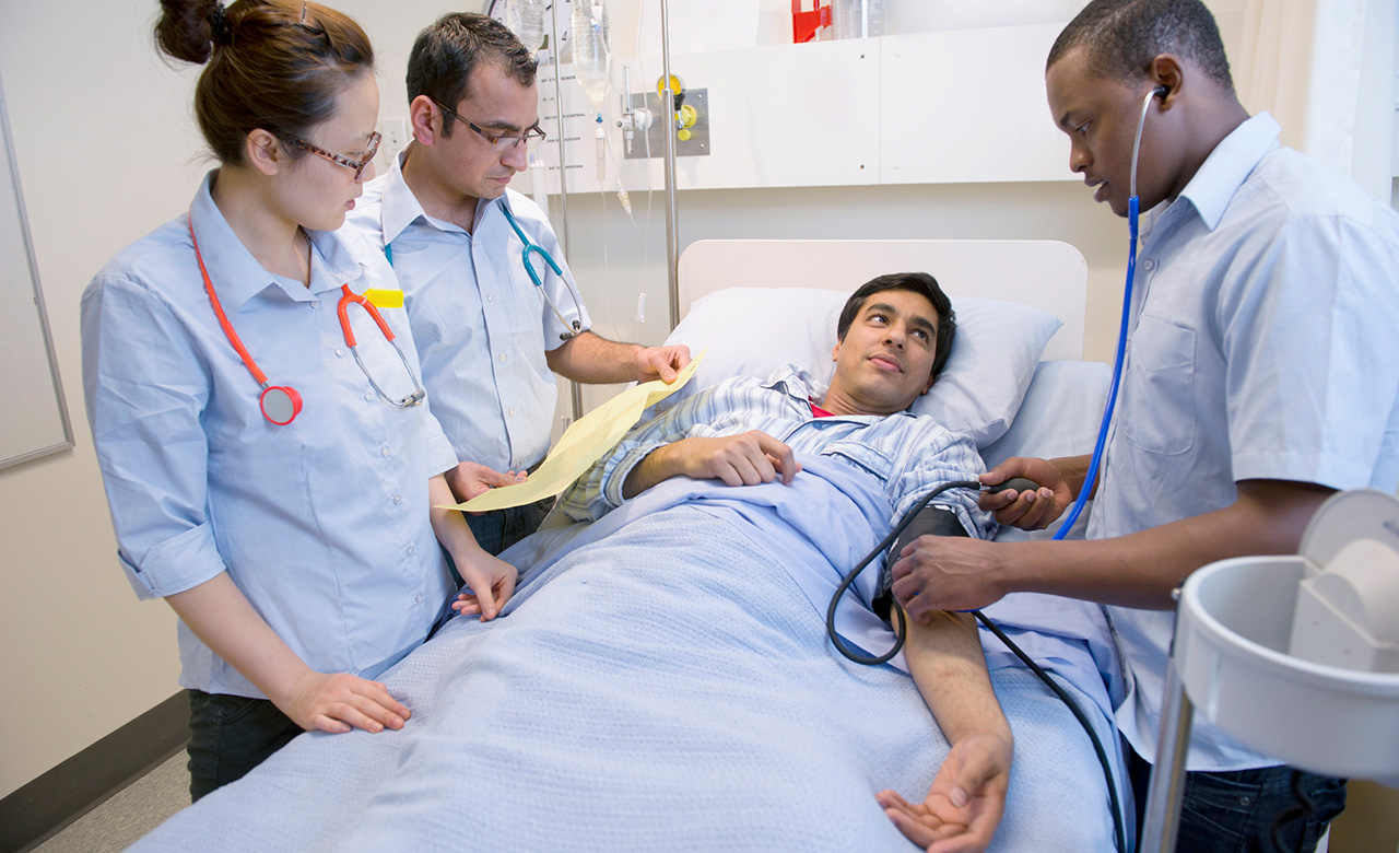 Four medical personnel surround a patient in a hospital bed. The patient's blood pressure is being measured with a blood pressure cuff and stethoscope. 