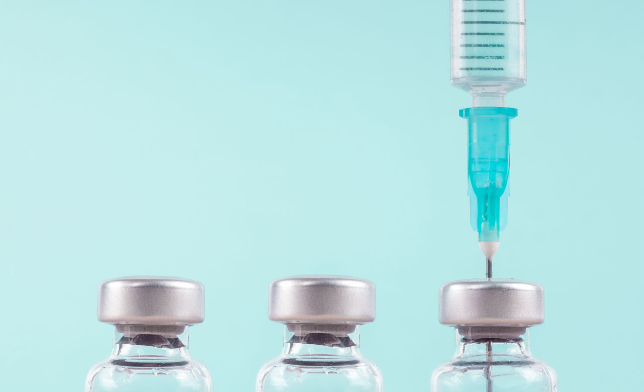 Three vials are lined up on a pale turquoise background, a syringe with a needle is inserted into the vial on the right. 