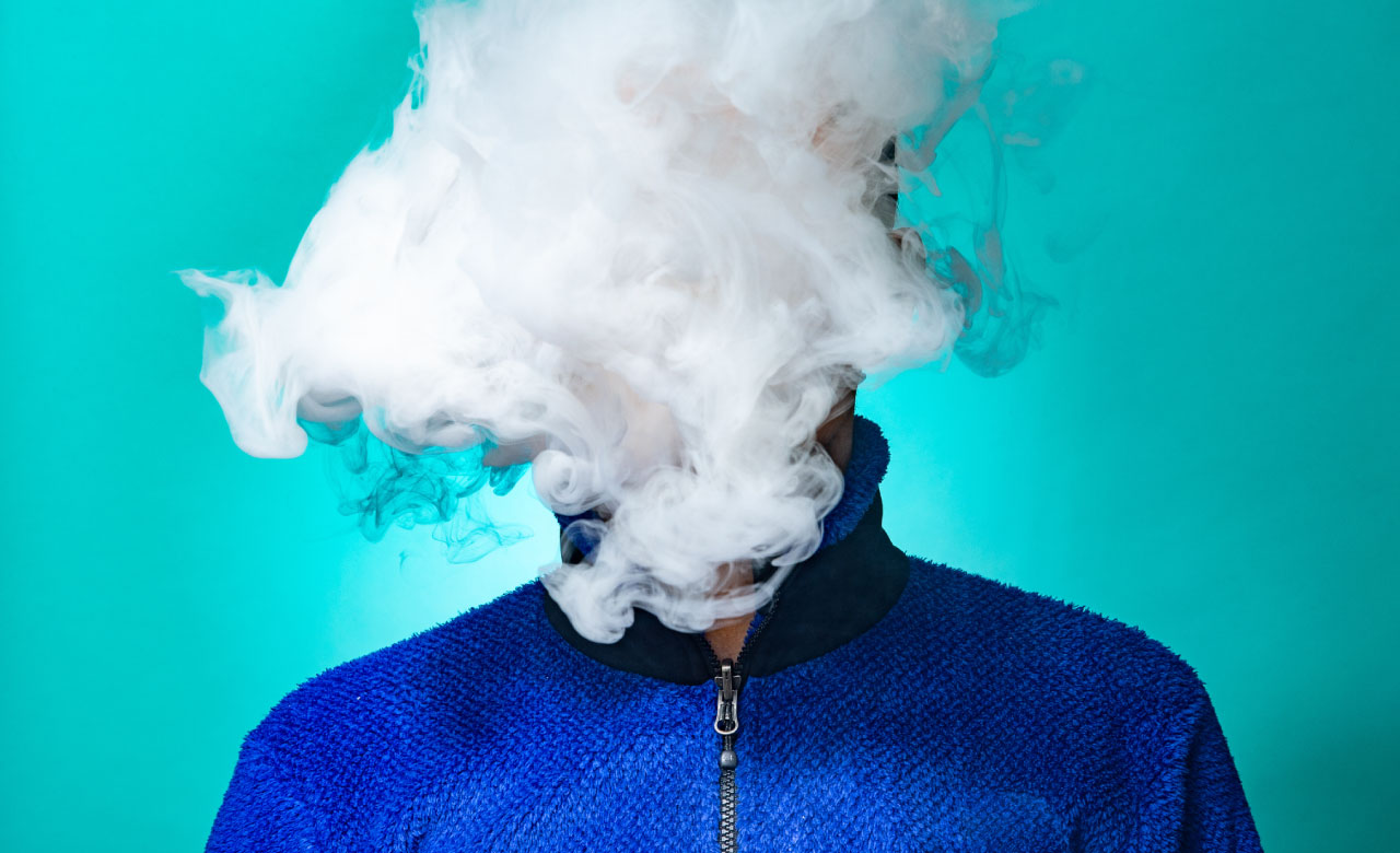 A person in a blue zip up jacket on a bright turquoise background with their head completely engulfed in a vape cloud