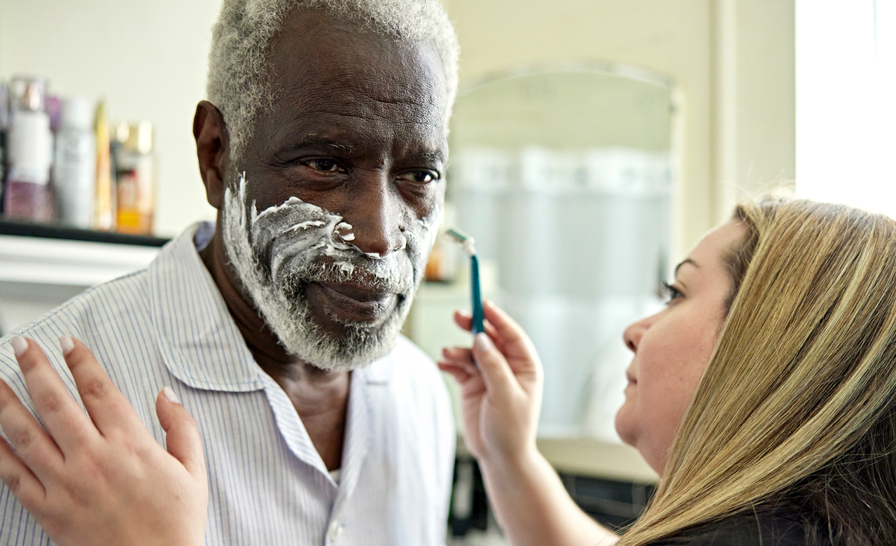 Alzheimer patient receiving assistance shaving from caregiver.